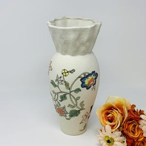 May include: Cream-colored ceramic vase with a textured surface and a ruffled top. The vase is adorned with colorful floral patterns in blue, yellow, and orange, with green leaves and stems. A decorative piece for home decor.
