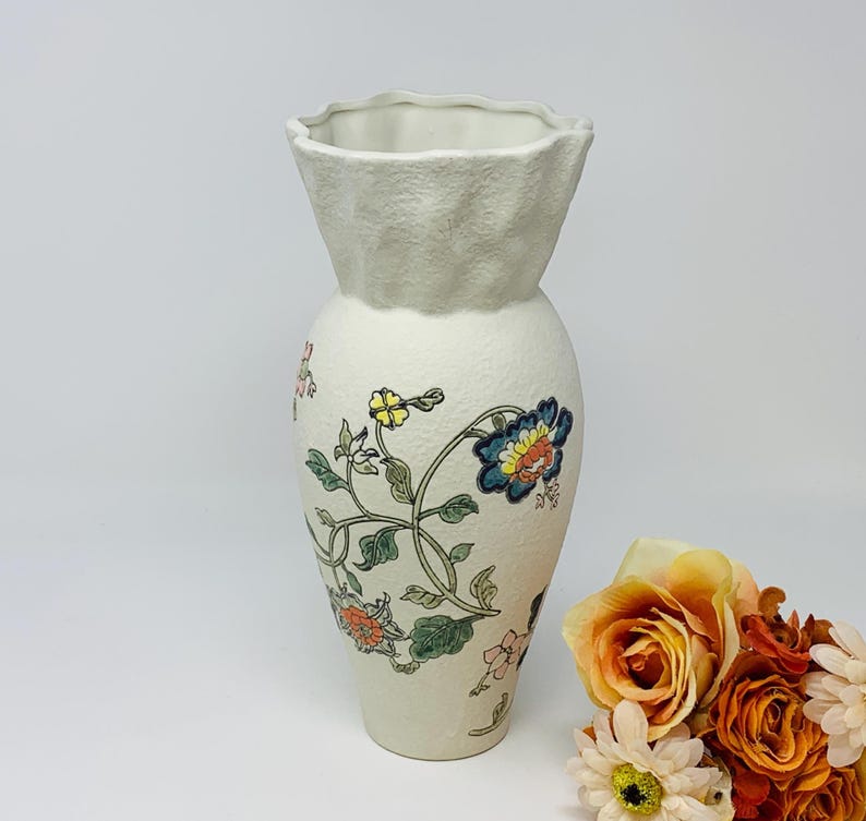 May include: Cream-colored ceramic vase with a textured surface and a ruffled top. The vase is adorned with colorful floral patterns in blue, yellow, and orange, with green leaves and stems. A decorative piece for home decor.