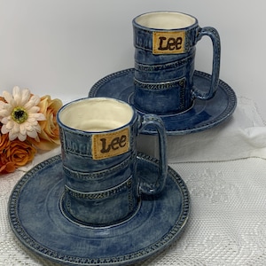 Puede incluir: Dos tazas y platillos de cerámica con diseño de mezclilla. Las tazas tienen asa y la palabra "Lee" en un parche. Los platillos y las tazas son azules con interior blanco. Un ramo de flores está en el fondo.