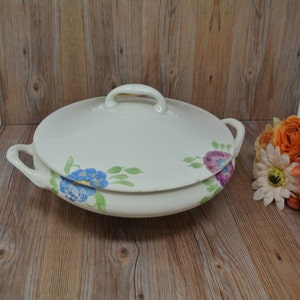 May include: White ceramic serving dish with a lid, featuring a floral design with blue, purple, and yellow flowers. The dish has two handles on the sides.