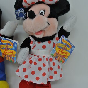 K&ouml;nnte beinhalten: Eine Pl&uuml;sch-Minnie-Maus-Puppe, die ein wei&szlig;es Kleid mit roten Punkten und eine rote Schleife tr&auml;gt. Die Puppe h&auml;lt ihre rechte Hand hoch und hat ein Etikett mit der Aufschrift "Der Zauber von Disney".