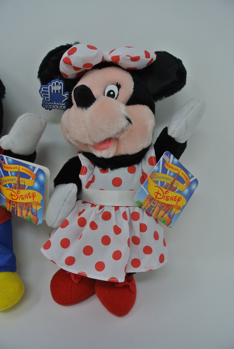 K&ouml;nnte beinhalten: Eine Pl&uuml;sch-Minnie-Maus-Puppe, die ein wei&szlig;es Kleid mit roten Punkten und eine rote Schleife tr&auml;gt. Die Puppe h&auml;lt ihre rechte Hand hoch und hat ein Etikett mit der Aufschrift "Der Zauber von Disney".