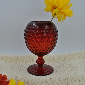 以下が含まれることがあります： 丸いボディと台座ベースのルビーレッドのホブネイルガラスの花瓶。明るい黄色の花が花瓶に入れられています。花瓶はクリーム色のレースのテーブルクロスの上にあります。