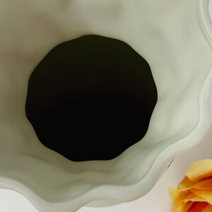 May include: A cream-colored ceramic vase with a wavy, scalloped rim. The interior is black, and a yellow rose is visible in the corner. The vase has a matte finish and a unique, organic shape.