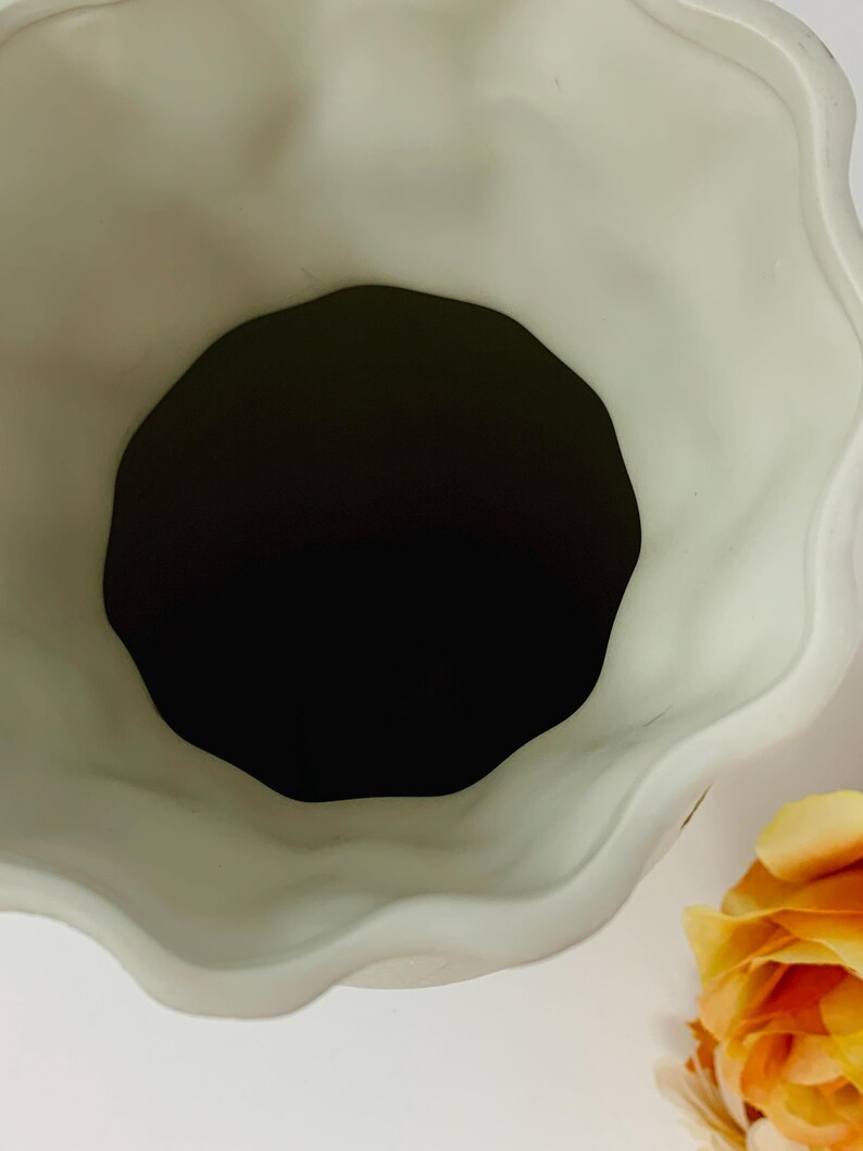 May include: A cream-colored ceramic vase with a wavy, scalloped rim. The interior is black, and a yellow rose is visible in the corner. The vase has a matte finish and a unique, organic shape.