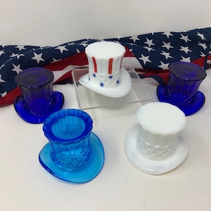 Könnte beinhalten: Eine Sammlung kleiner, zylinderförmiger Glasbehälter in verschiedenen Farben, darunter Blau und Weiß. Ein Hut hat rot-blaue Streifen und Sterne, mit einer amerikanischen Flagge im Hintergrund. Diese dekorativen Artikel sind wahrscheinlich für patriotische Feiertage gedacht.