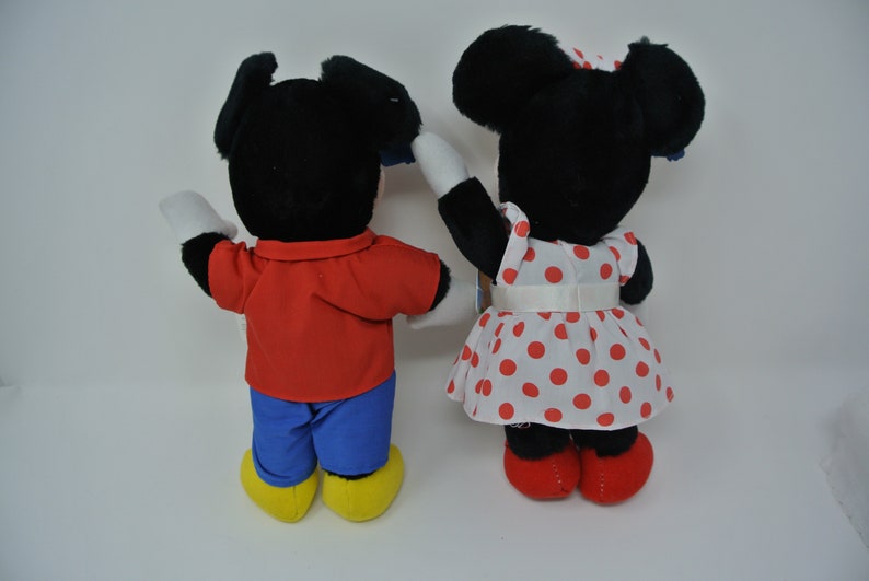 K&ouml;nnte beinhalten: Ein Paar Pl&uuml;schtiere, Mickey Mouse und Minnie Mouse, stehen R&uuml;cken an R&uuml;cken. Mickey tr&auml;gt ein rotes Hemd und eine blaue Hose, w&auml;hrend Minnie ein wei&szlig;es Kleid mit roten Punkten und einer roten Schleife tr&auml;gt.