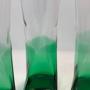 Op de afbeelding: Drie heldere, hoge glazen met een groene gradi&euml;nt aan de basis. De glazen hebben een unieke, licht gebogen vorm. Het woord "Tanqueray" is in het wit op de bodem van elk glas gedrukt. De glazen zijn waarschijnlijk bedoeld voor het serveren van cocktails of drankjes.