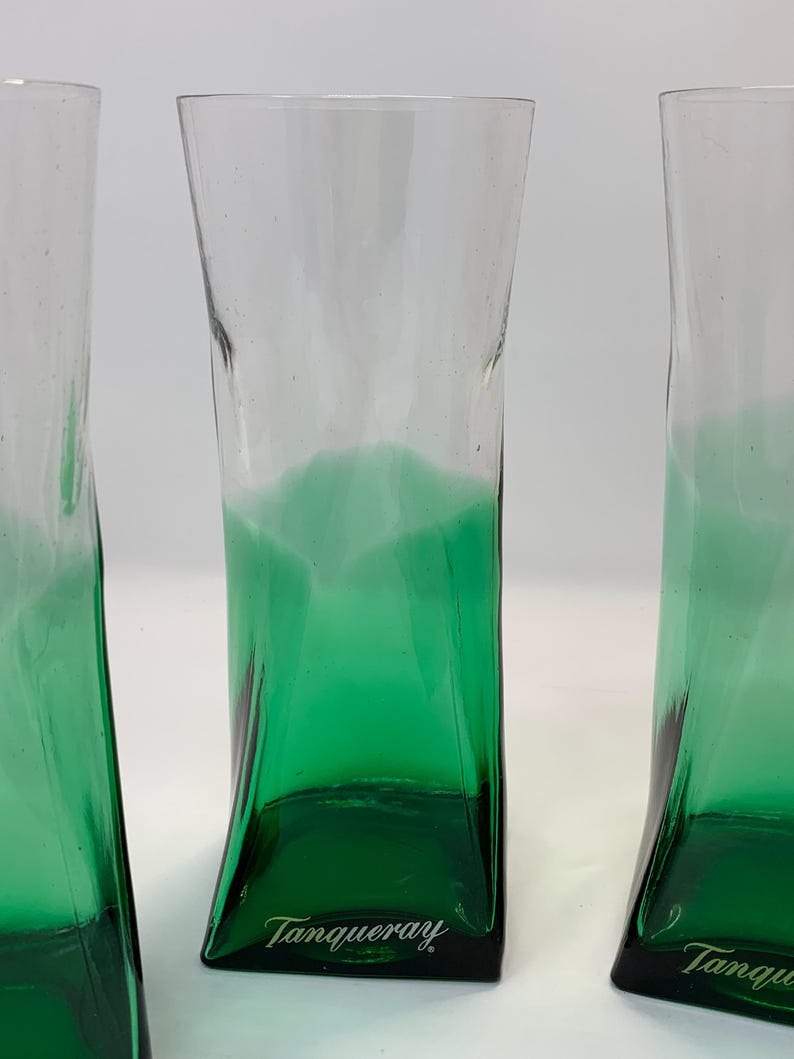 Op de afbeelding: Drie heldere, hoge glazen met een groene gradi&euml;nt aan de basis. De glazen hebben een unieke, licht gebogen vorm. Het woord "Tanqueray" is in het wit op de bodem van elk glas gedrukt. De glazen zijn waarschijnlijk bedoeld voor het serveren van cocktails of drankjes.
