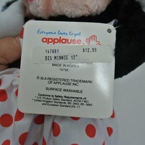 K&ouml;nnte beinhalten: Ein wei&szlig;es Pl&uuml;sch-Minnie-Maus-Spielzeug mit roten Punkten und einer schwarzen Schleife. Das Spielzeug ist 30,5 cm gro&szlig; und hat ein Etikett mit der Aufschrift "Everyone loves to get applause" und "DIS MINNIE 12" mit einem Preis von 12,99 $. Auf dem Etikett steht auch "Made in Korea" und "Oberfl&auml;che waschbar".