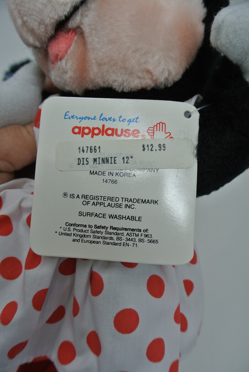 K&ouml;nnte beinhalten: Ein wei&szlig;es Pl&uuml;sch-Minnie-Maus-Spielzeug mit roten Punkten und einer schwarzen Schleife. Das Spielzeug ist 30,5 cm gro&szlig; und hat ein Etikett mit der Aufschrift "Everyone loves to get applause" und "DIS MINNIE 12" mit einem Preis von 12,99 $. Auf dem Etikett steht auch "Made in Korea" und "Oberfl&auml;che waschbar".