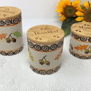 May include: Three ceramic canisters with wooden lids, decorated with fall leaves and acorns. The lids are engraved with the words "Traditions", "Thankful", and "Harvest". The canisters have a cream-colored base with a brown and orange floral border.