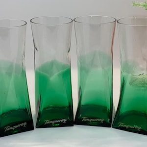 Op de afbeelding: Vier transparante glazen met een groene tint en een uniek, hoekig ontwerp. De basis van elk glas is donkergroen, overgaand in transparant aan de bovenkant. Het woord "Tanqueray" is op de basis van elk glas gedrukt.