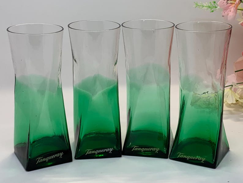 Op de afbeelding: Vier transparante glazen met een groene tint en een uniek, hoekig ontwerp. De basis van elk glas is donkergroen, overgaand in transparant aan de bovenkant. Het woord "Tanqueray" is op de basis van elk glas gedrukt.