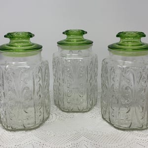 Op de afbeelding: Drie heldere glazen potten met groene deksels. De potten hebben een vierkante vorm met reliëf bloempatronen. De deksels zijn felgroen van kleur. De potten worden gepresenteerd op een wit kanten tafelkleed.
