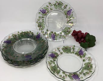 May include: A set of clear glass plates with scalloped edges, each featuring a hand-painted design of purple grapes and green leaves around the rim. A cluster of artificial red grapes with green leaves is positioned next to the plates.