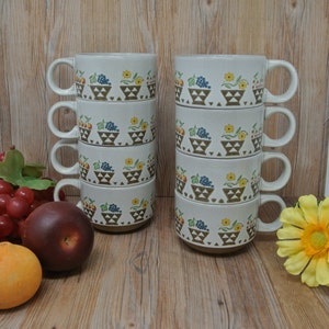 Può includere: Set di sei tazze in ceramica bianca con un motivo a ceste di frutta marrone, giallo, verde e blu. Le tazze sono impilate in due gruppi da tre.