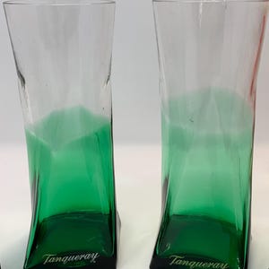 Op de afbeelding: Twee heldere, hoge glazen met een groene basis en een gebogen ontwerp. De basis van elk glas is donkergroen, met het woord "Tanqueray" in het wit gedrukt. De glazen zijn geschikt voor cocktails of andere dranken.