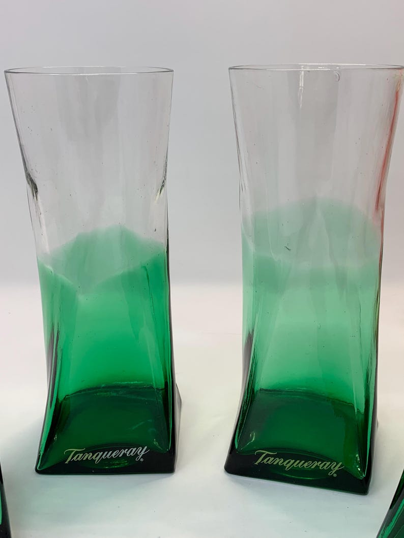 Op de afbeelding: Twee heldere, hoge glazen met een groene basis en een gebogen ontwerp. De basis van elk glas is donkergroen, met het woord "Tanqueray" in het wit gedrukt. De glazen zijn geschikt voor cocktails of andere dranken.
