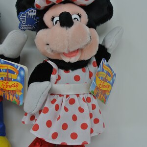 K&ouml;nnte beinhalten: Ein Pl&uuml;schtier von Minnie Mouse, das ein wei&szlig;es Kleid mit roten Punkten und eine rote Schleife tr&auml;gt. Das Spielzeug hat eine rosa Nase und schwarze Augen. Das Spielzeug h&auml;lt ein Schild mit der Aufschrift "Jeder liebt einen guten Applaus!" und ein weiteres Schild mit der Aufschrift "Die Feier von Disney".