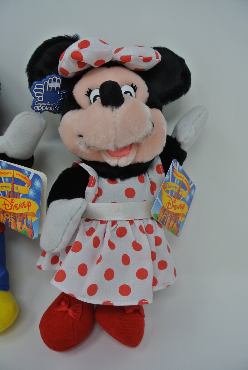 K&ouml;nnte beinhalten: Ein Pl&uuml;schtier von Minnie Mouse, das ein wei&szlig;es Kleid mit roten Punkten und eine rote Schleife tr&auml;gt. Das Spielzeug hat eine rosa Nase und schwarze Augen. Das Spielzeug h&auml;lt ein Schild mit der Aufschrift "Jeder liebt einen guten Applaus!" und ein weiteres Schild mit der Aufschrift "Die Feier von Disney".