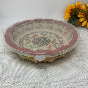 May include: A round ceramic baking dish with a decorative fall-themed pattern of leaves and acorns. The dish has a red rim and a clear plastic lid. The dish is sitting on a white lace tablecloth, with sunflowers in the background.