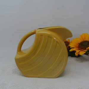 Puede incluir: Jarra de cerámica de color amarillo mostaza con asa curva y boquilla. La jarra presenta una serie de líneas verticales y acanaladas en su cuerpo redondeado. Se exhibe sobre un mantel de encaje blanco, con girasoles en el fondo.
