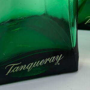 Op de afbeelding: Close-up van een groene glazen fles met het woord "Tanqueray" in een cursief lettertype. De fles heeft een donkergroene basis en een helder, reflecterend oppervlak. De afbeelding benadrukt het logo van het merk.