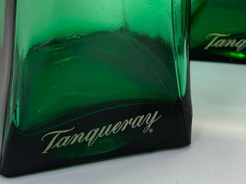 Op de afbeelding: Close-up van een groene glazen fles met het woord "Tanqueray" in een cursief lettertype. De fles heeft een donkergroene basis en een helder, reflecterend oppervlak. De afbeelding benadrukt het logo van het merk.