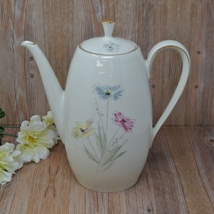 Puede incluir: Una cafetera de cerámica blanca con ribete dorado y diseño floral. Las flores son rosas, azules y amarillas.