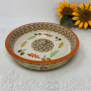 Puede incluir: Fuente redonda de cerámica para pastel con borde naranja e interior crema. La fuente presenta un patrón floral marrón alrededor del borde y un diseño central de flores marrones. Hojas y bellotas coloridas están dispersas en el interior.