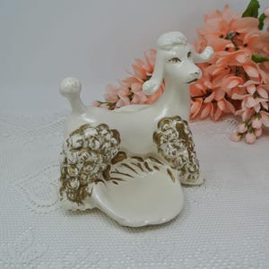 Puede incluir: Figura de caniche vintage de cerámica blanca con detalles dorados. El caniche tiene un pelaje decorativo y texturizado y un pequeño cuenco delante. La figura es un objeto decorativo, posiblemente para guardar objetos pequeños.
