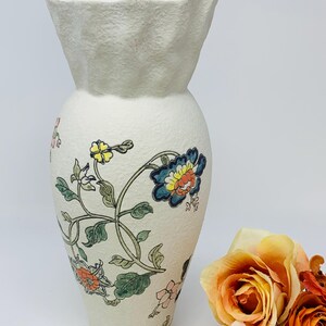 May include: A cream-colored ceramic vase with a textured surface and a flared, ruffled top. The vase is adorned with a floral design featuring blue, orange, and yellow flowers with green leaves. The vase is approximately 10 inches tall.
