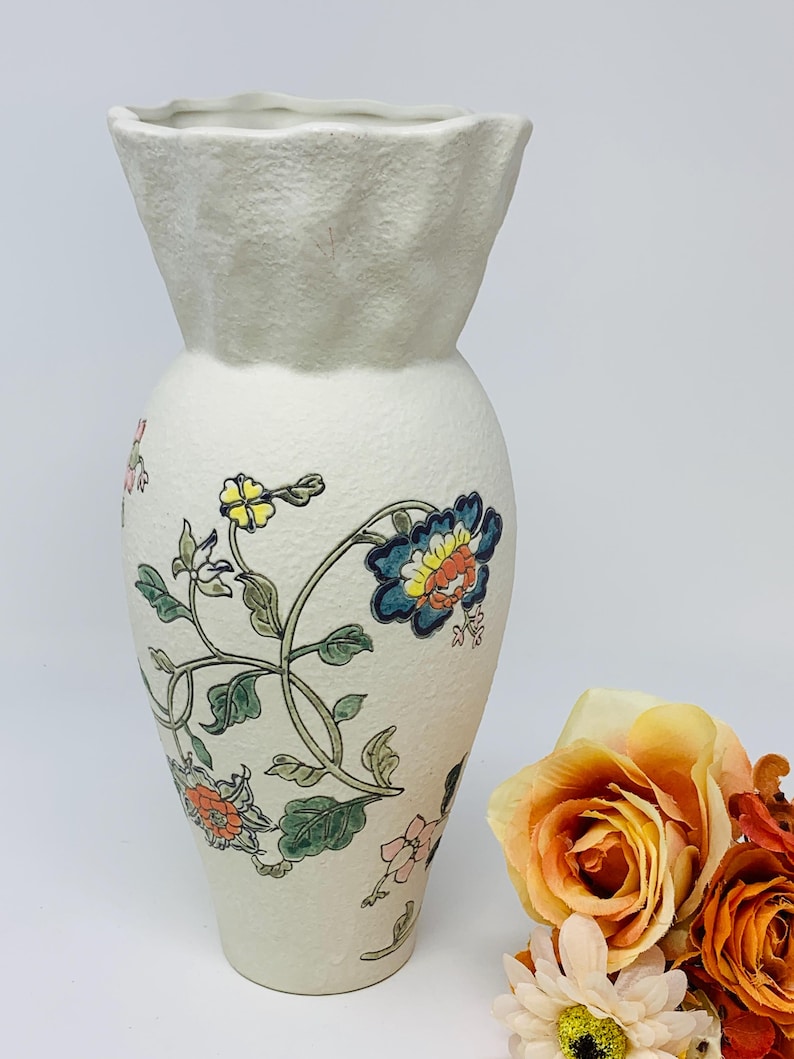 May include: A cream-colored ceramic vase with a textured surface and a flared, ruffled top. The vase is adorned with a floral design featuring blue, orange, and yellow flowers with green leaves. The vase is approximately 10 inches tall.