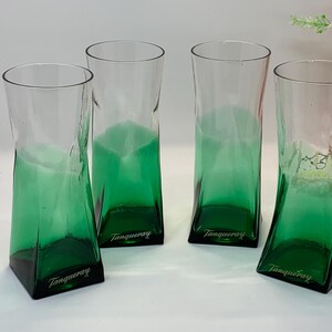 Op de afbeelding: Vier heldere glazen bekers met een groene basis en een licht gedraaid ontwerp. Elk glas heeft het woord "Tanqueray" op de basis gedrukt. De glazen zijn gerangschikt op een wit oppervlak.