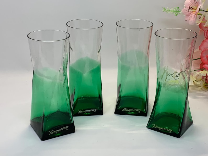 Op de afbeelding: Vier heldere glazen bekers met een groene basis en een licht gedraaid ontwerp. Elk glas heeft het woord "Tanqueray" op de basis gedrukt. De glazen zijn gerangschikt op een wit oppervlak.