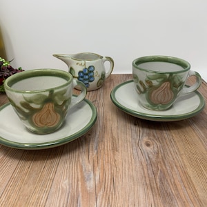 Könnte beinhalten: Zwei Keramik-Teetassen und Untertassen mit einem grünen und braunen Birnenmuster. Ein passender Milchkännchen mit einem Traubenmuster ist ebenfalls abgebildet.