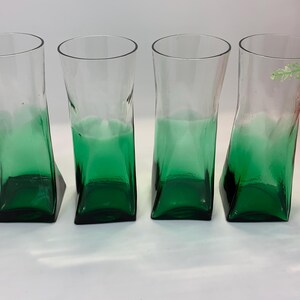 Op de afbeelding: Vier heldere glazen bekers met een groene basis en een uniek, gebogen ontwerp. De glazen hebben een vierkante basis en een licht gekantelde, rechthoekige body. De groene kleur vervaagt naar boven, waardoor een ombre-effect ontstaat. De glazen zijn geschikt voor het serveren van dranken.