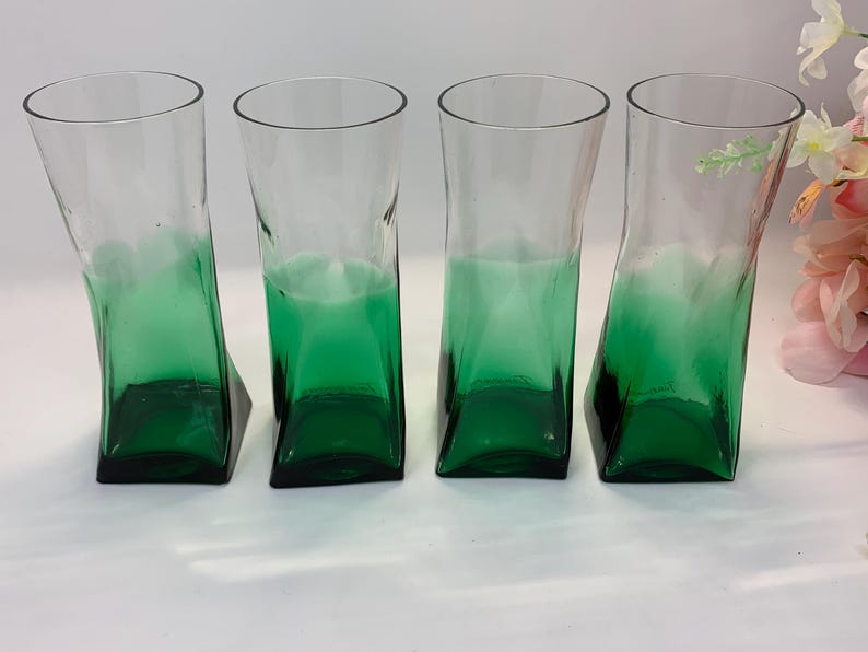 Op de afbeelding: Vier heldere glazen bekers met een groene basis en een uniek, gebogen ontwerp. De glazen hebben een vierkante basis en een licht gekantelde, rechthoekige body. De groene kleur vervaagt naar boven, waardoor een ombre-effect ontstaat. De glazen zijn geschikt voor het serveren van dranken.