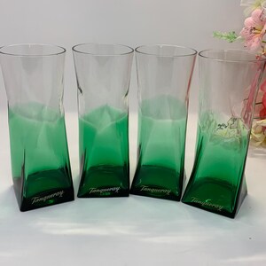 Op de afbeelding: Vier transparante glazen met een groene tint en een opvallende, schuine vorm. De basis van elk glas is effen donkergroen, met het woord "Tanqueray" erop gedrukt. Deze glazen zijn geschikt voor het serveren van cocktails of andere dranken.