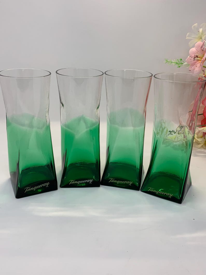 Op de afbeelding: Vier transparante glazen met een groene tint en een opvallende, schuine vorm. De basis van elk glas is effen donkergroen, met het woord "Tanqueray" erop gedrukt. Deze glazen zijn geschikt voor het serveren van cocktails of andere dranken.