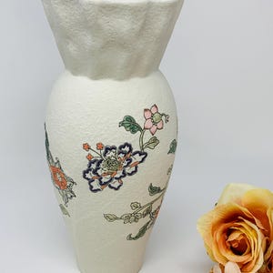 May include: Off-white ceramic vase with a textured surface and a flared top. The vase is adorned with colorful floral patterns in shades of orange, blue, and pink. A decorative piece for home decor.