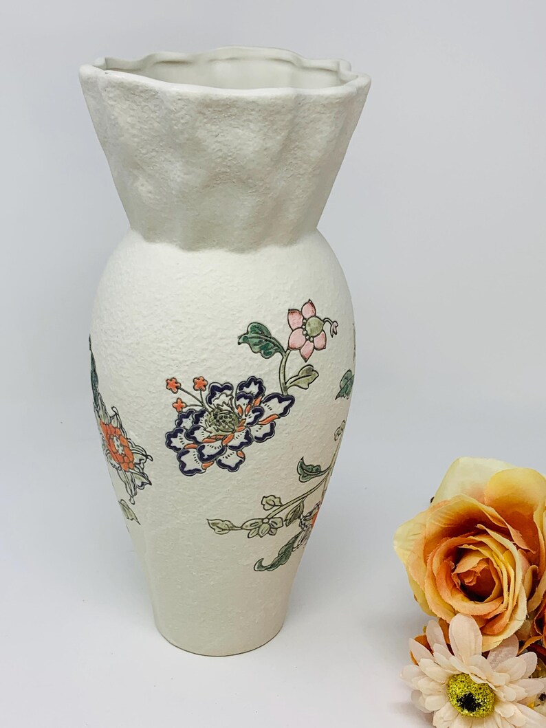 May include: Off-white ceramic vase with a textured surface and a flared top. The vase is adorned with colorful floral patterns in shades of orange, blue, and pink. A decorative piece for home decor.