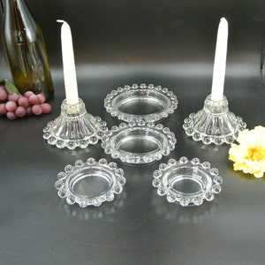 Pode incluir: Conjunto de cinco castiçais de vidro transparente com bordas peroladas. Os castiçais são de tamanhos diferentes e estão dispostos em fila sobre uma superfície preta.