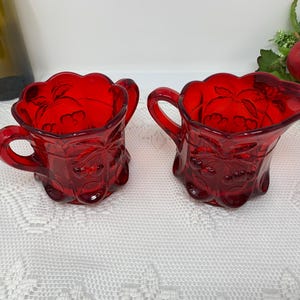 Può includere: Due lattine di vetro rosso rubino con manici. Le lattine hanno un motivo floreale e un bordo smerlato. Le lattine sono su una tovaglia di pizzo bianco. Una bottiglia verde e della frutta sono sullo sfondo.