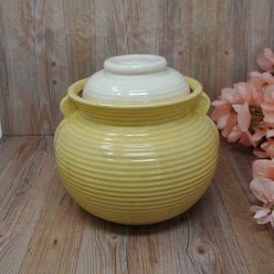 May include: A yellow ceramic jar with a lid. The jar has a ribbed design and two small handles. The lid is white and round. The jar is sitting on a wooden surface.