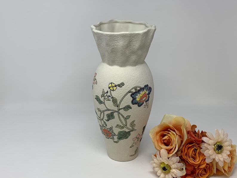 May include: A cream-colored ceramic vase with a textured surface and a ruffled top. The vase is adorned with floral patterns in shades of blue, orange, and green. A bouquet of orange and white flowers sits nearby.