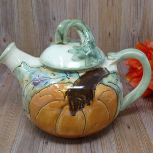 May include: A ceramic teapot with a pumpkin design. The teapot features a light green handle and lid, with a pumpkin design in shades of orange and brown. The spout is light cream colored, and the teapot has a whimsical, hand-painted style.