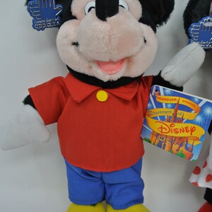 K&ouml;nnte beinhalten: Ein Pl&uuml;sch-Mickey-Mouse-Spielzeug, das ein rotes Hemd, eine blaue Hose und gelbe Schuhe tr&auml;gt. Das Spielzeug h&auml;lt ein blaues Etikett mit der Aufschrift "Jeder liebt Applaus". Das Spielzeug steht auf einer wei&szlig;en Oberfl&auml;che. Das Etikett auf dem Spielzeug lautet "Applause pr&auml;sentiert den Zauber von Disney".