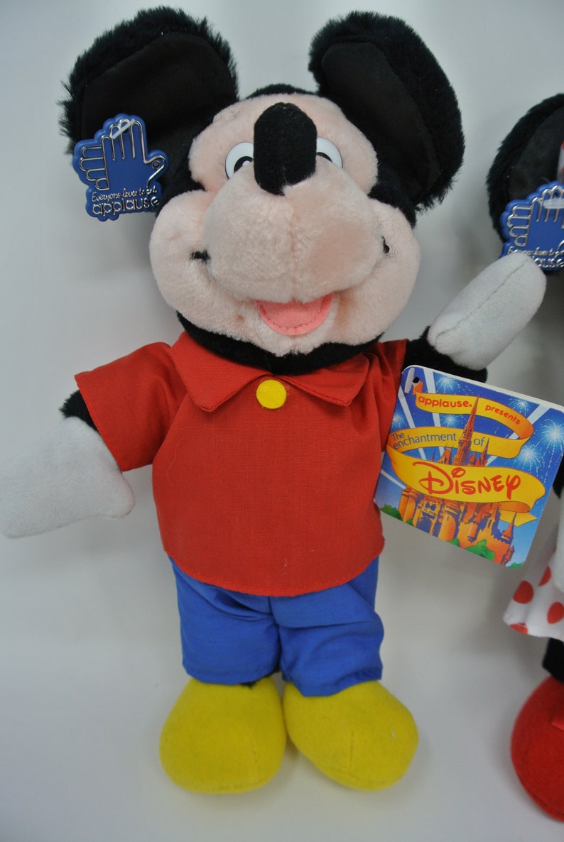 K&ouml;nnte beinhalten: Ein Pl&uuml;sch-Mickey-Mouse-Spielzeug, das ein rotes Hemd, eine blaue Hose und gelbe Schuhe tr&auml;gt. Das Spielzeug h&auml;lt ein blaues Etikett mit der Aufschrift "Jeder liebt Applaus". Das Spielzeug steht auf einer wei&szlig;en Oberfl&auml;che. Das Etikett auf dem Spielzeug lautet "Applause pr&auml;sentiert den Zauber von Disney".